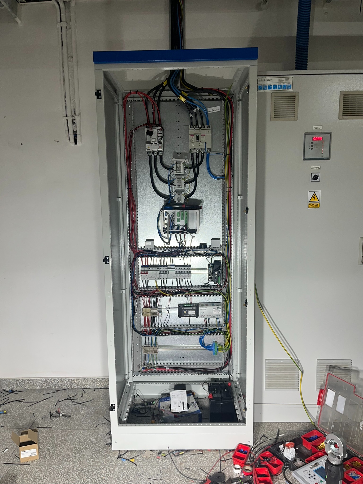 Otwarta szafa elektryczna z widocznymi przewodami, bezpiecznikami i elementami sterującymi. Na podłodze narzędzia i fragmenty przewodów.