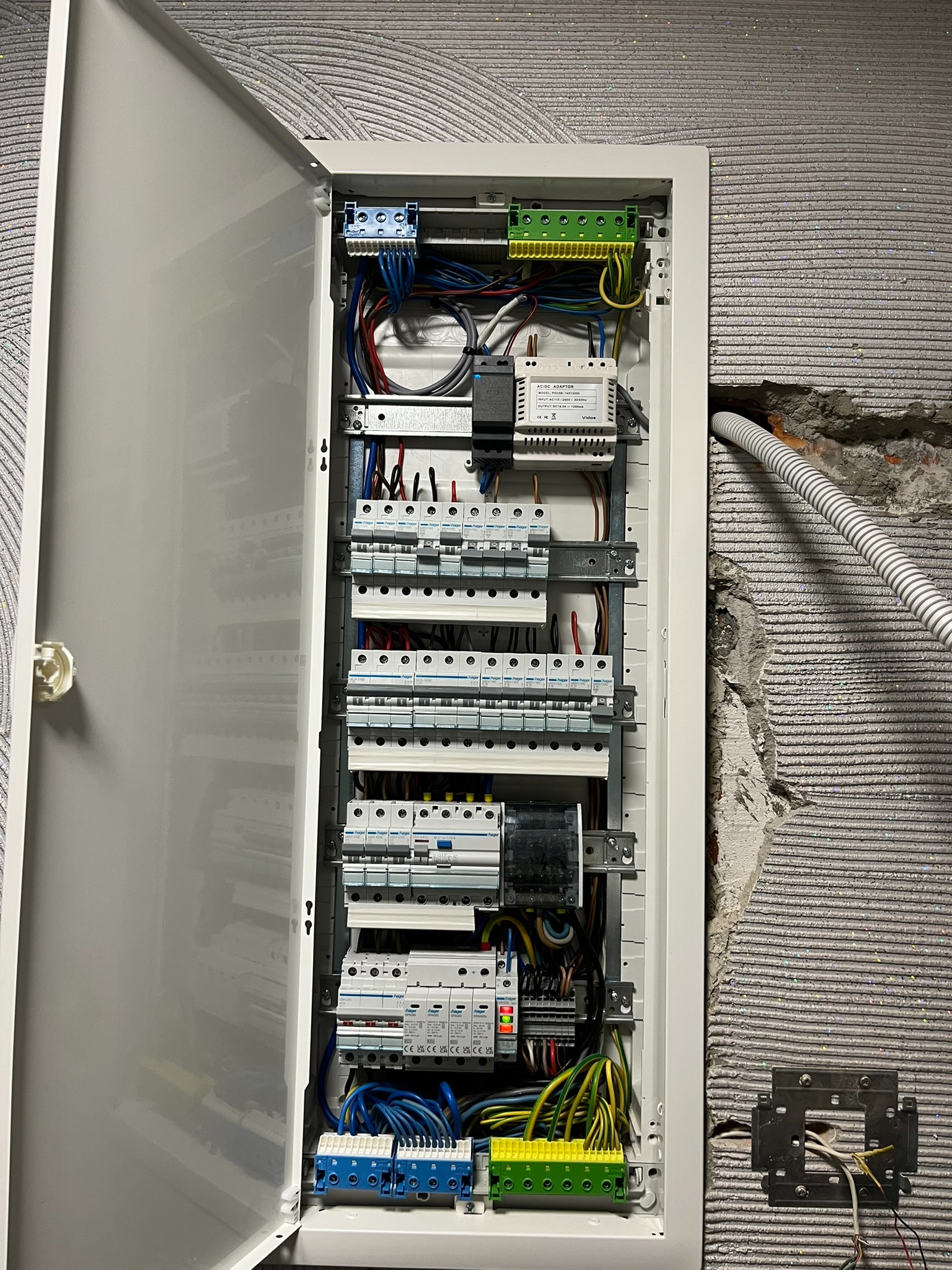 Otwarta skrzynka elektryczna z widocznymi bezpiecznikami i przewodami na tle ściany z szorstką fakturą. Widoczny fragment instalacji w ścianie.