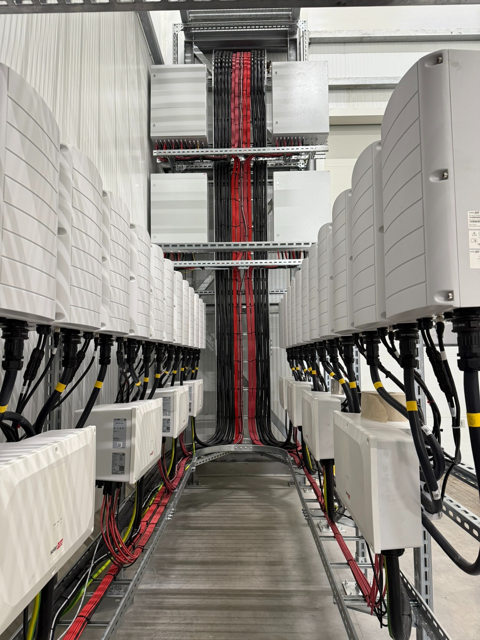 Rząd białych skrzynek elektrycznych z czarnymi i czerwonymi kablami na metalowych szynach, widok z perspektywy w hali z blachy falistej.