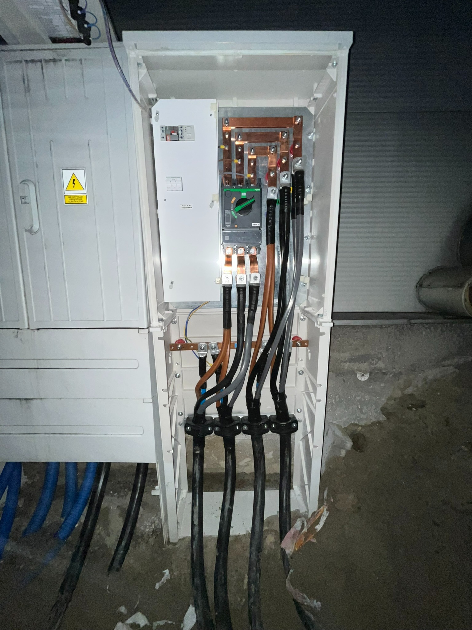 Rozdzielnica elektryczna z podłączonymi kablami zasilającymi. Widoczne zabezpieczenia i przewody w skrzynce. Instalacja w trakcie prac wykończeniowych.