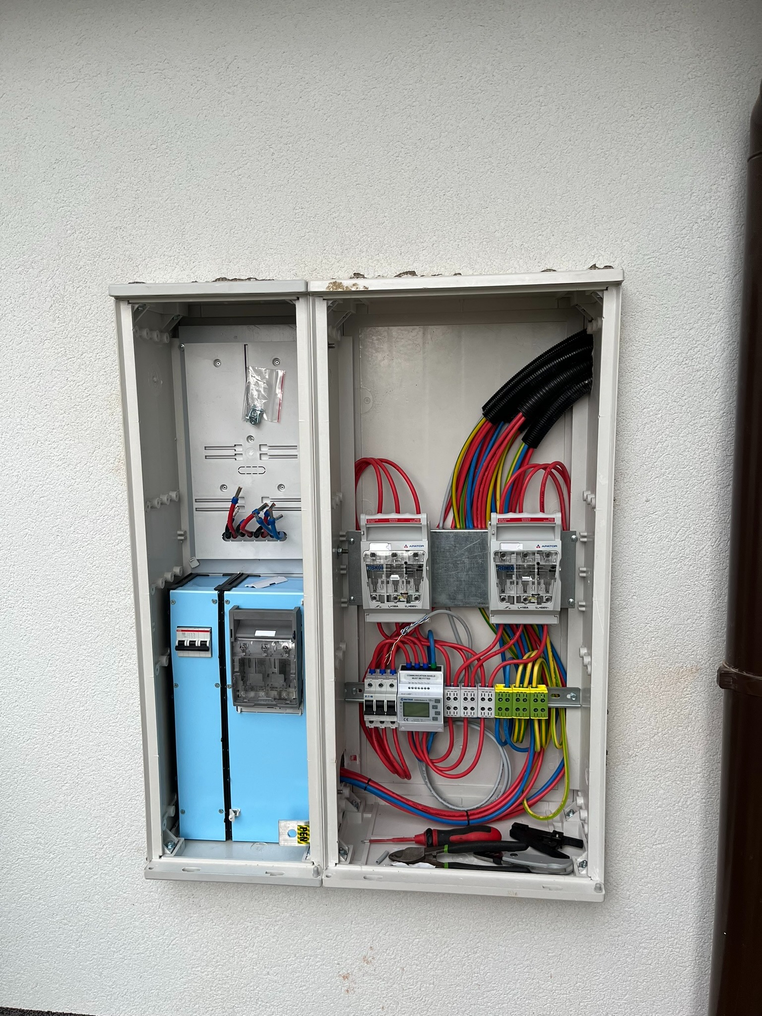 Otwarta skrzynka elektryczna na białej ścianie, z widocznymi przewodami, licznikami, bezpiecznikami i narzędziami, sugerująca instalację lub naprawę elektryczną.
