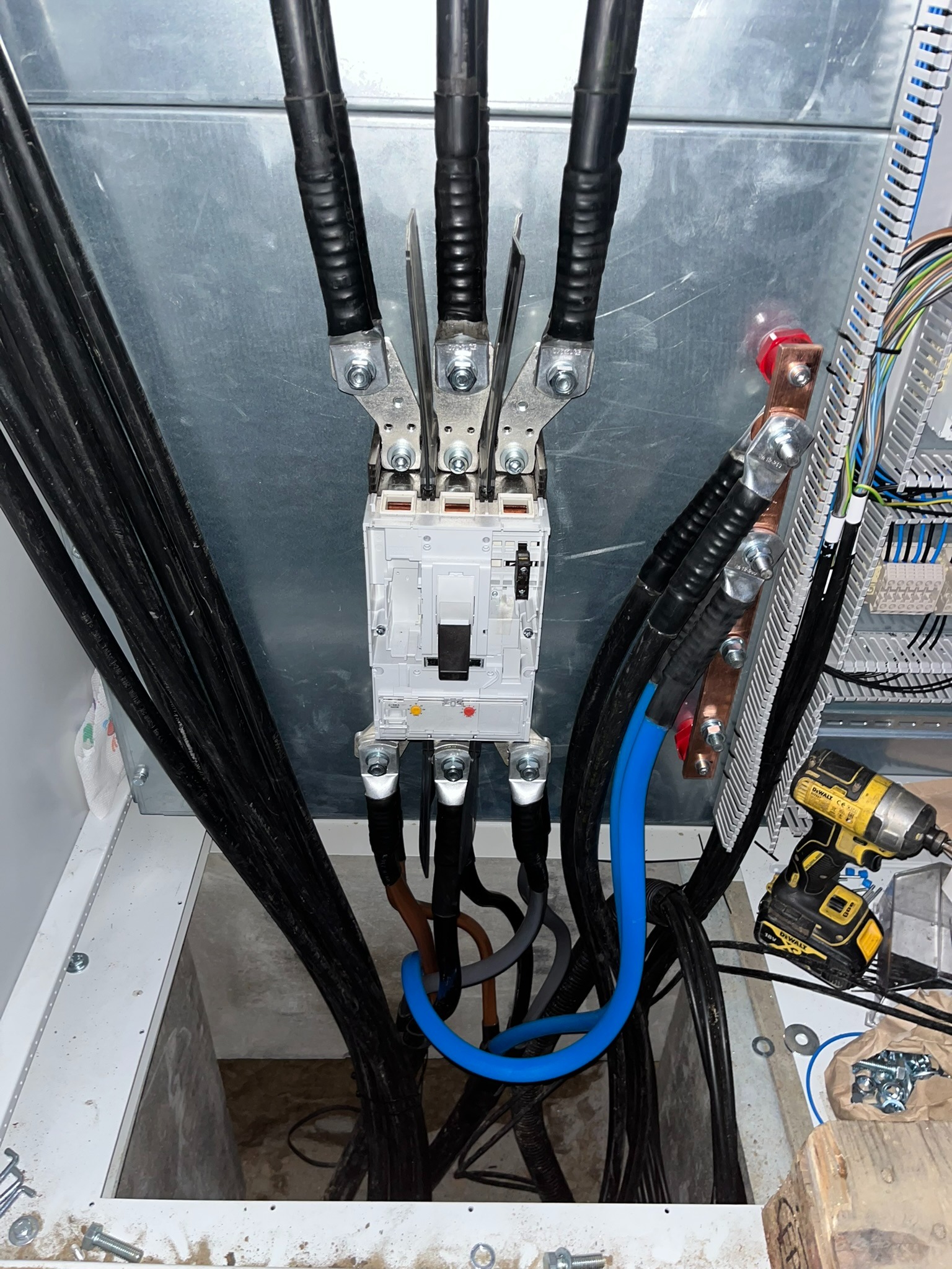 Szczegółowe ujęcie z góry na podłączone kable elektryczne do rozdzielnicy z użyciem srebrnych złączek. Widoczny niebieski kabel, wiertarka i śruby.