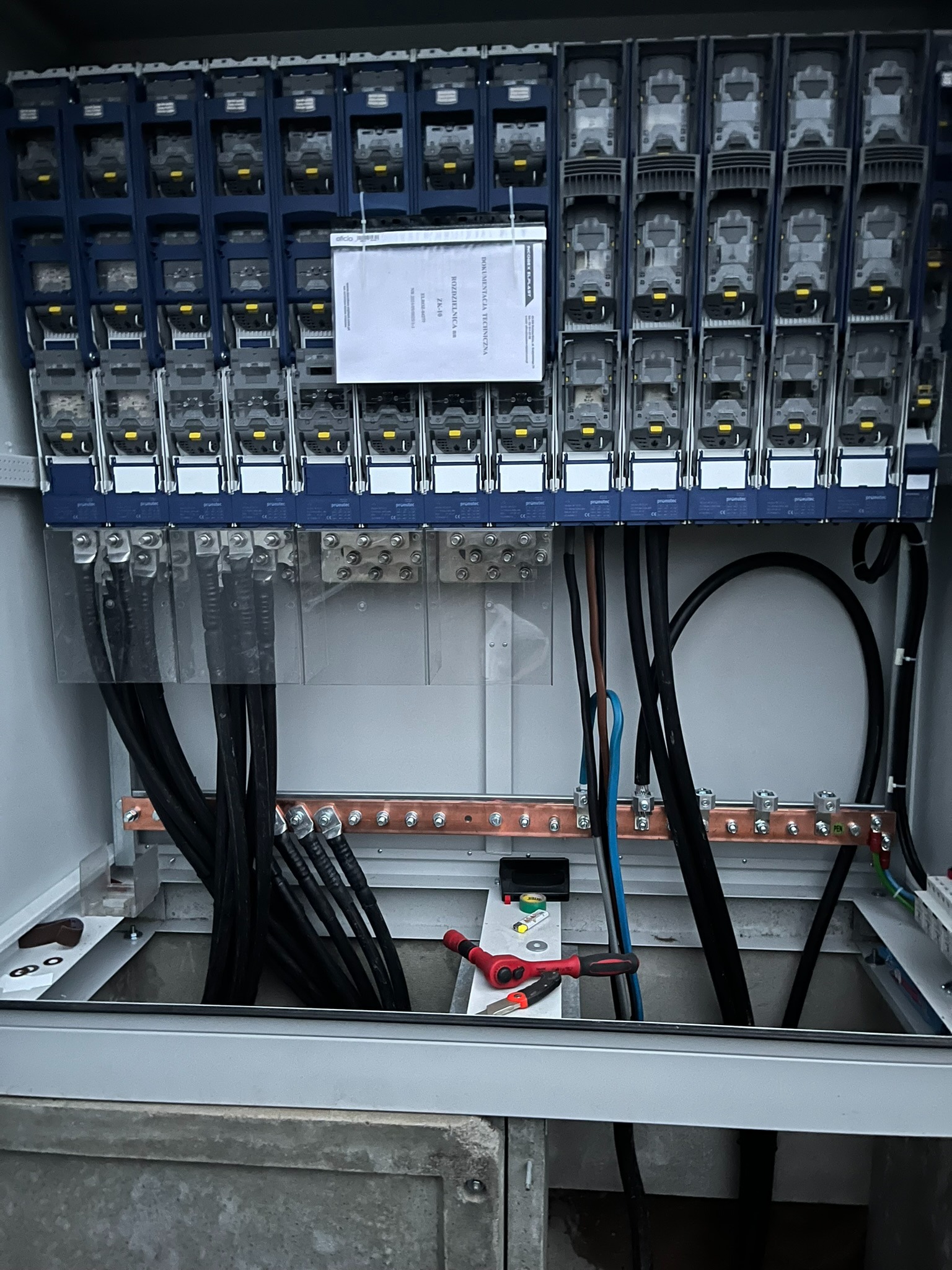 Rozdzielnia elektryczna z zamontowanymi bezpiecznikami, widoczne kable, listwy zaciskowe i narzędzia. Tabliczka z dokumentacją techniczną na górze.