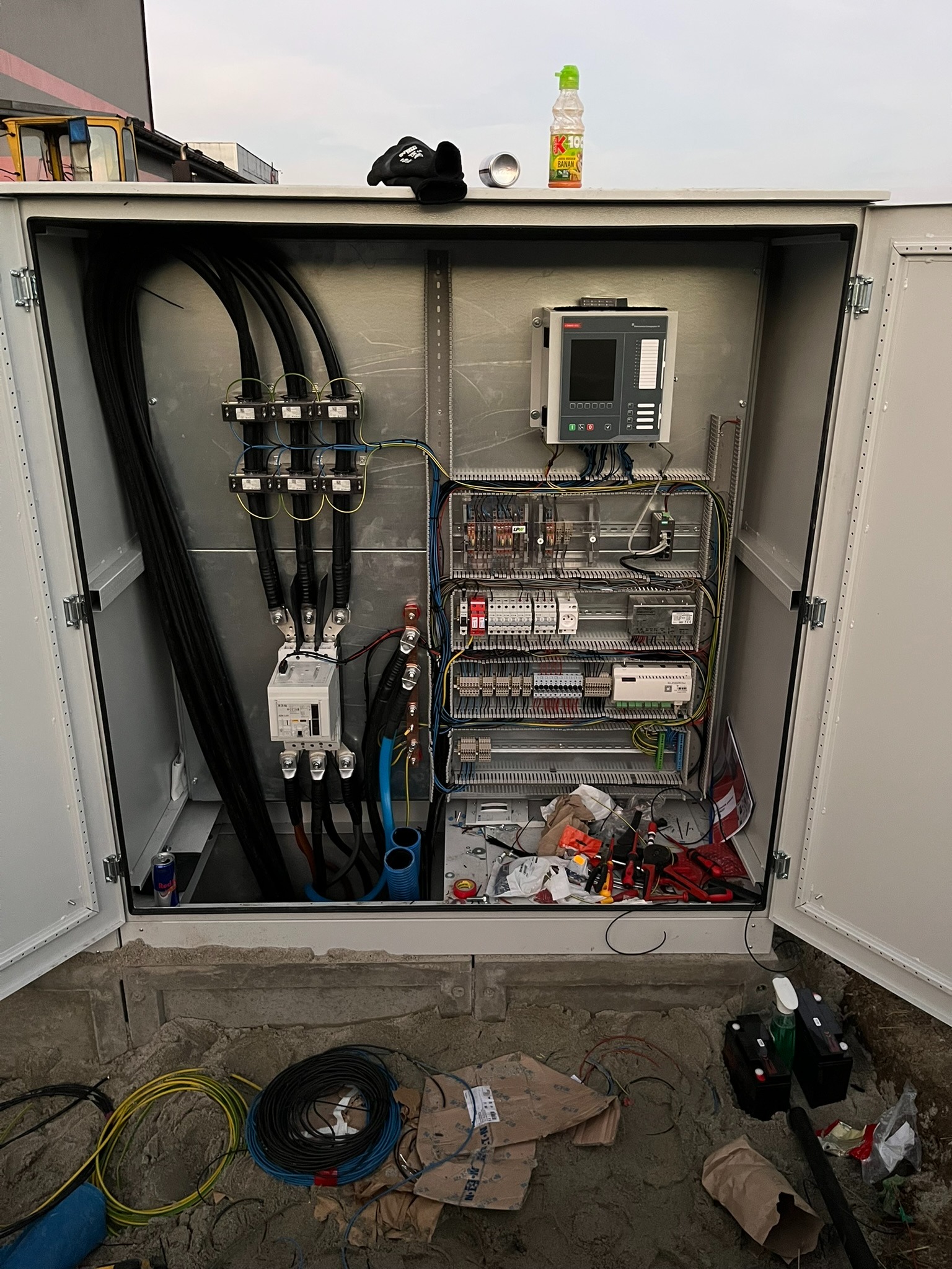 Otwarta skrzynka elektryczna z zainstalowaną aparaturą i kablami. Na górze butelka soku i rękawice. Na dole kable i narzędzia. Realizacja instalacji elektrycznej.