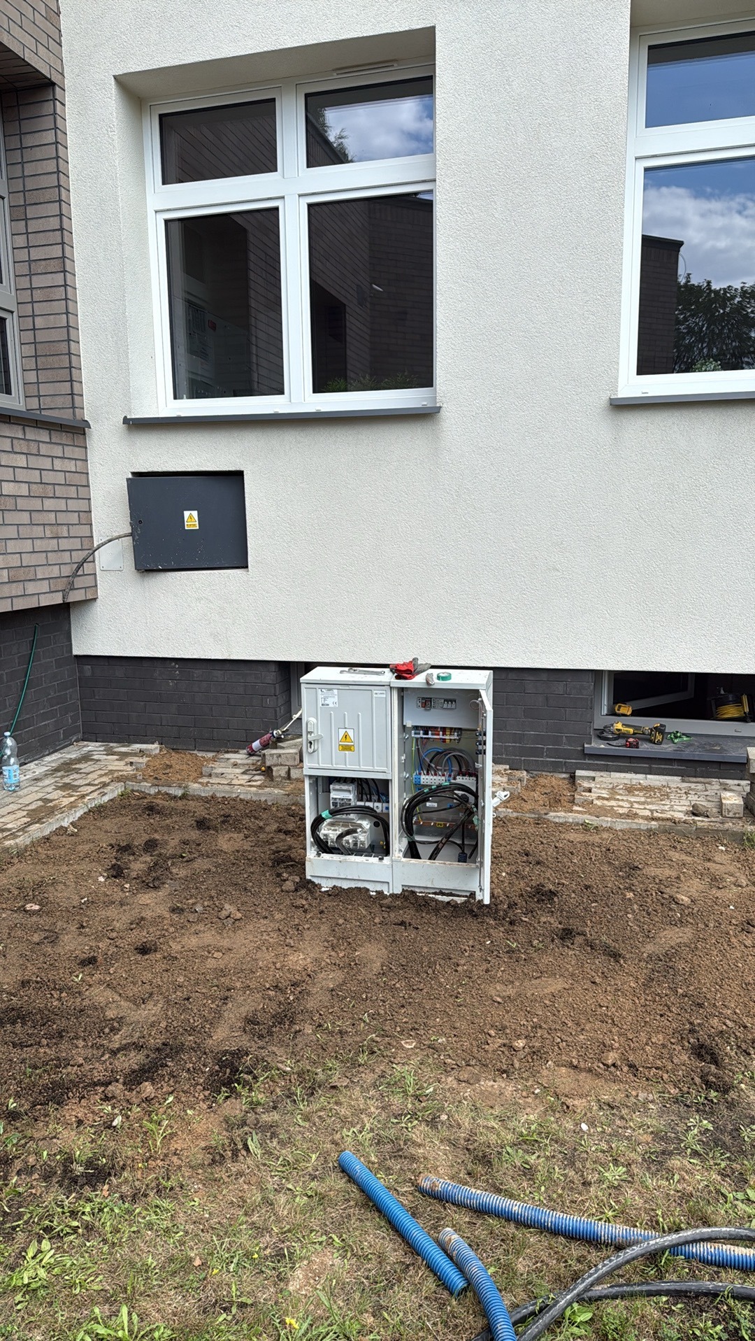 Otwarta skrzynka elektryczna na zewnątrz budynku, widoczne kable i bezpieczniki, narzędzia obok, w tle okna budynku i fragment muru z cegły.