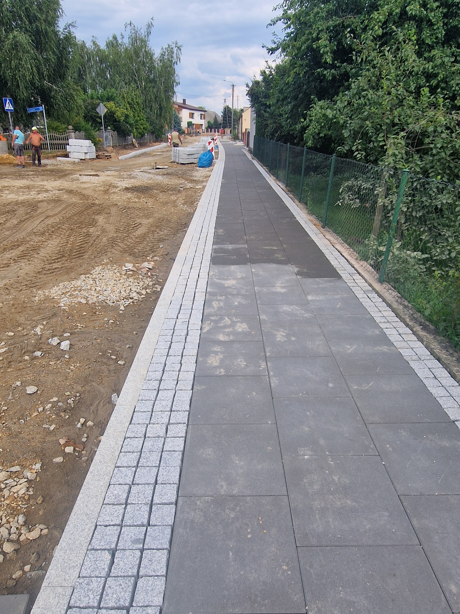 Fragment nowo wyłożonego chodnika z szarych płyt betonowych obramowanych kostką granitową, z widocznym placem budowy w tle i pracownikami w oddali.