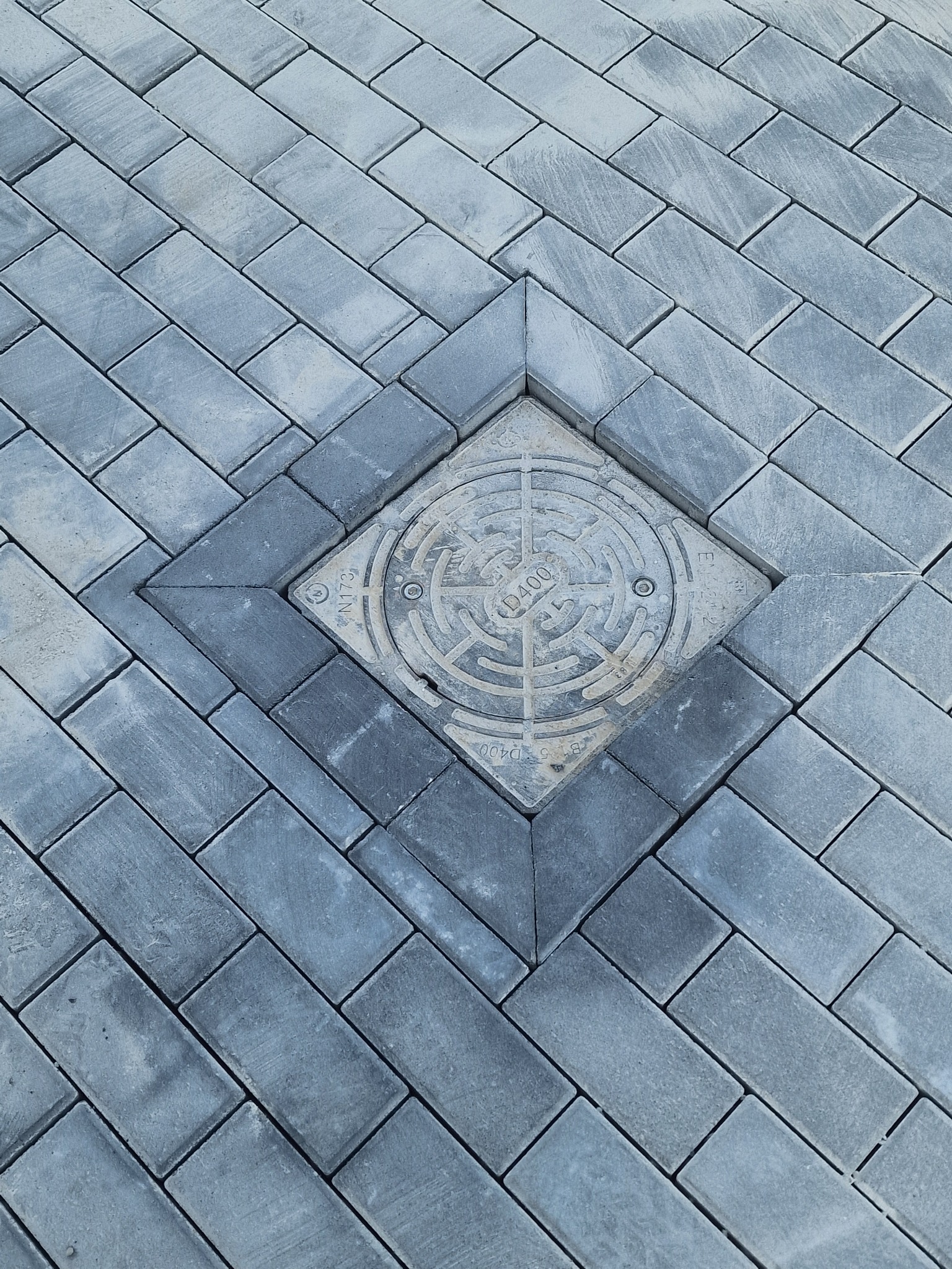 Szara kostka brukowa ułożona w jodełkę z kwadratową studzienką kanalizacyjną pośrodku.