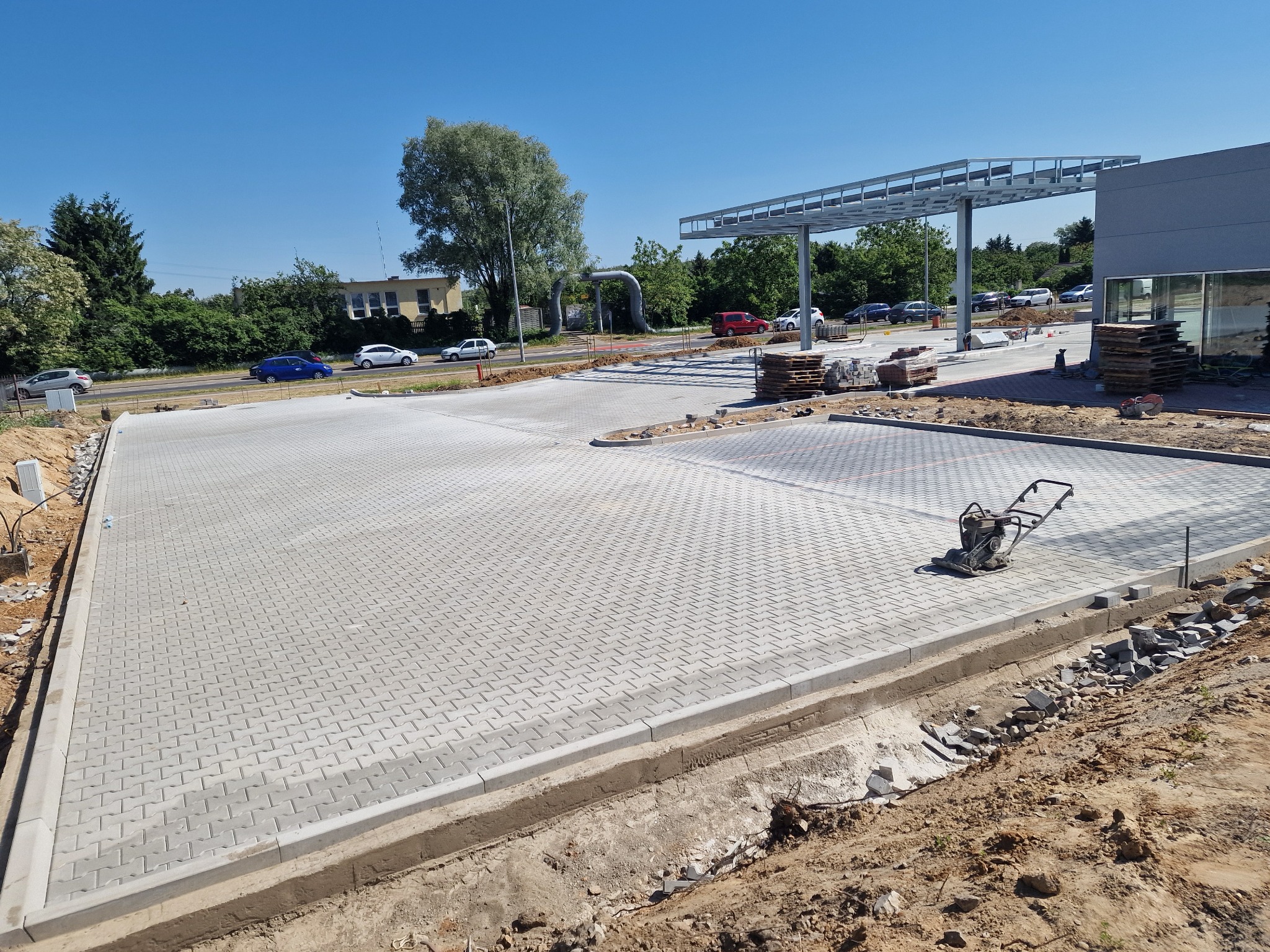 Rozległy plac wyłożony szarą kostką brukową w trakcie budowy, z widocznym zagęszczarką i przygotowanym obrzeżem, na tle konstrukcji zadaszenia i zieleni.