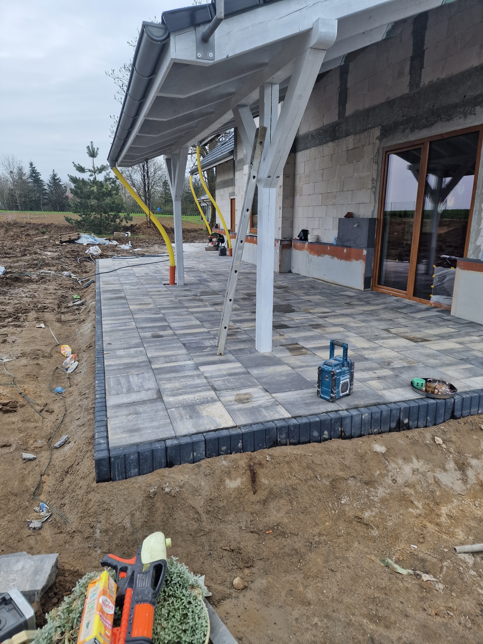Nowo ułożony taras z szarej kostki brukowej, obrzeża z ciemnej kostki, widoczny fragment domu w trakcie budowy, biała pergola i drabina oparta o słup.