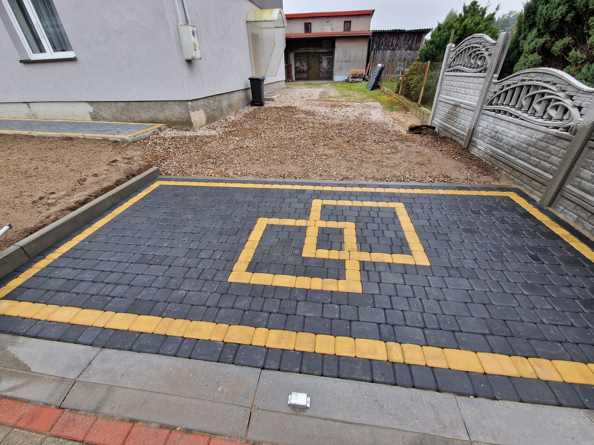 Ułożona nawierzchnia z czarno-żółtej kostki brukowej w kształcie dwóch przeplatających się kwadratów, widok z góry, w tle budynek i ogrodzenie z betonowych elementów.