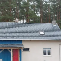 Gotowy dach z szarej dachówki ceramicznej z oknem dachowym i kamiennym kominem na tle lasu iglastego.