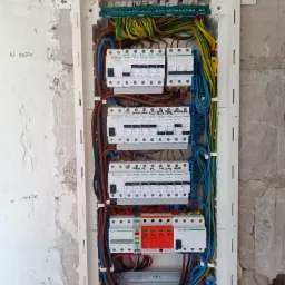 Rozdzielnica elektryczna w trakcie montażu, widoczne liczne przewody w różnych kolorach oraz zabezpieczenia marki Schneider, surowa ściana w tle z widocznymi śladami po tynku.