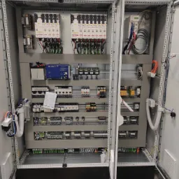 Otwarty panel sterowania z widocznymi elementami elektrycznymi, w tym sterowniki, zasilacze, przekaźniki i okablowanie. Widoczna dokumentacja techniczna wewnątrz szafy. Górna część obudowy...