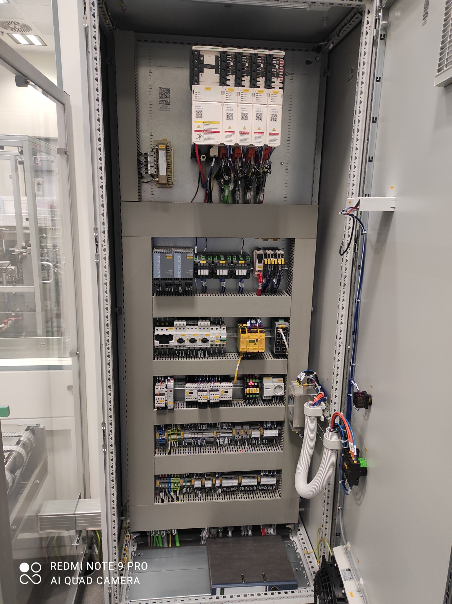 Otwarta szafa sterownicza z widocznymi modułami PLC, zasilaczami, bezpiecznikami i okablowaniem, zainstalowana w pomieszczeniu przemysłowym, widoczne elementy linii produkcyjnej za szybą.