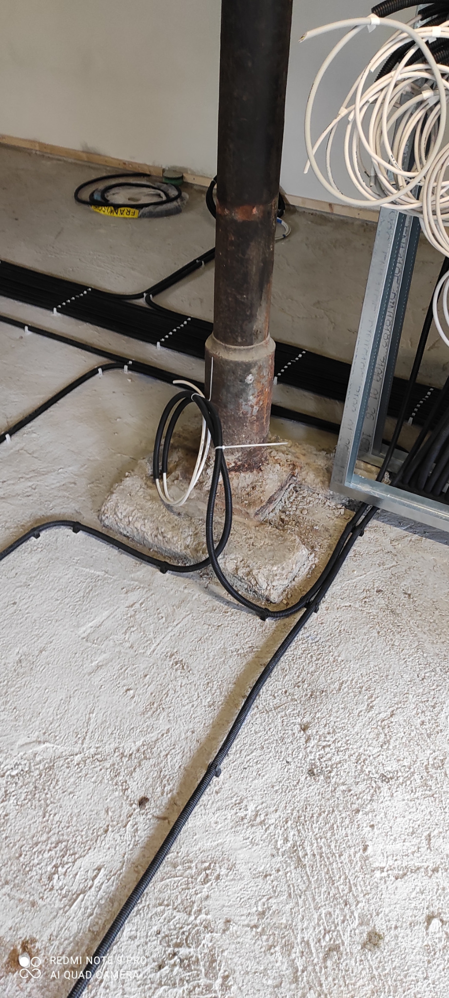 Surowe wnętrze w trakcie prac: czarne i białe kable elektryczne ułożone na surowym betonie wokół metalowej rury, widoczne mocowania kabli do podłoża.