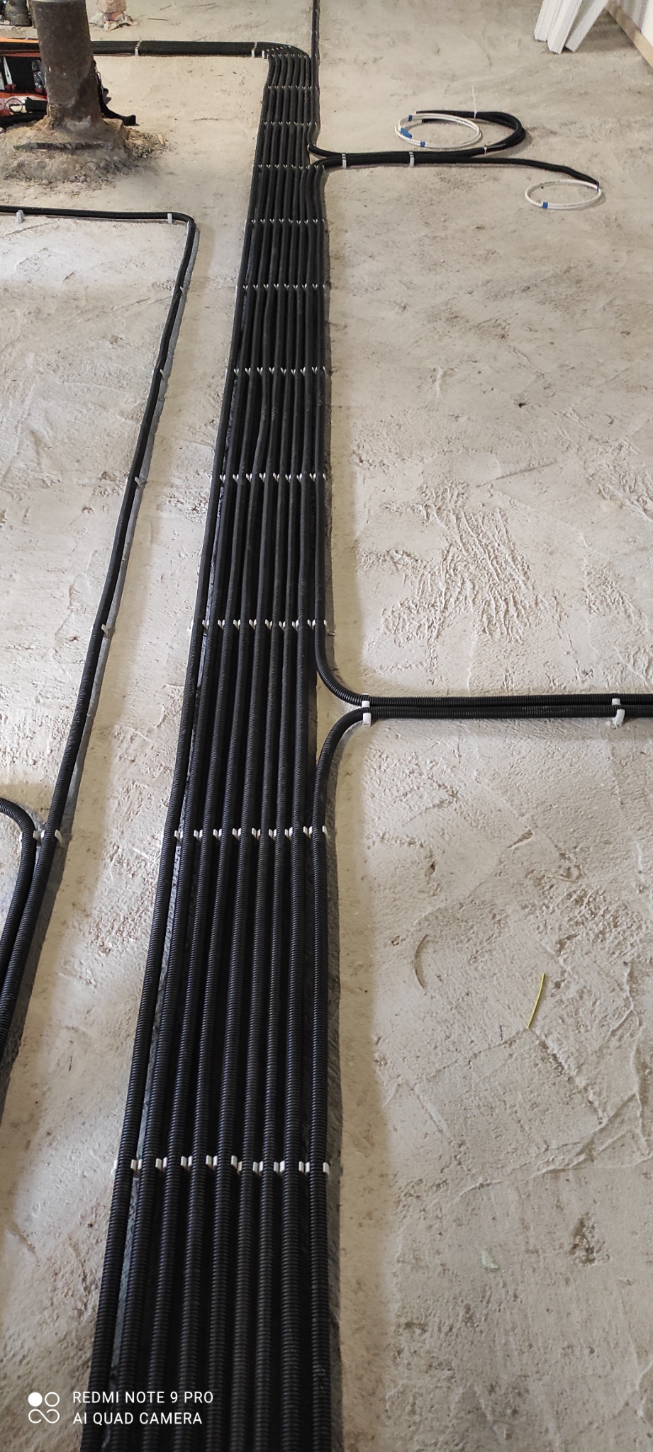 Równolegle ułożone, czarne rury karbowane z przewodami elektrycznymi, przymocowane do surowej, jasnej ściany za pomocą białych uchwytów. Widoczne fragmenty innych rur i przewodów, oraz surowy...