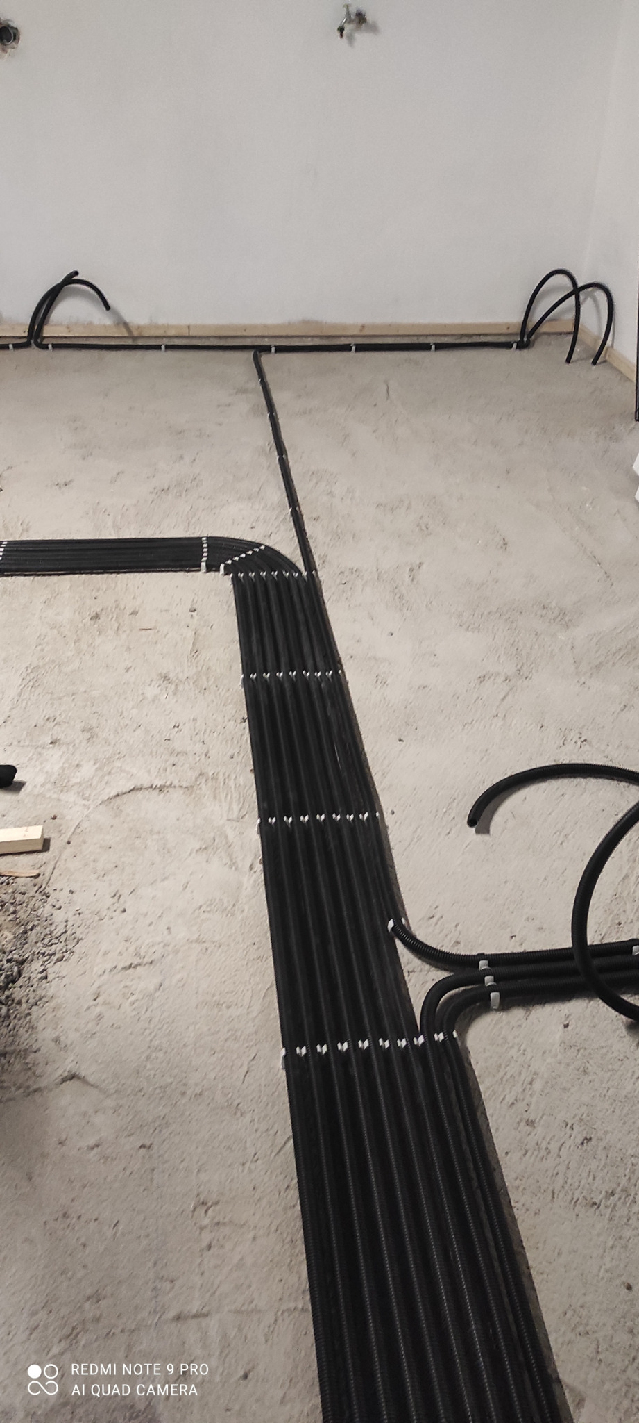 Czarny peszel ułożony równolegle, zamocowany do surowego betonu podłogi w Złotowie, w tle ściana z wystającymi przewodami.