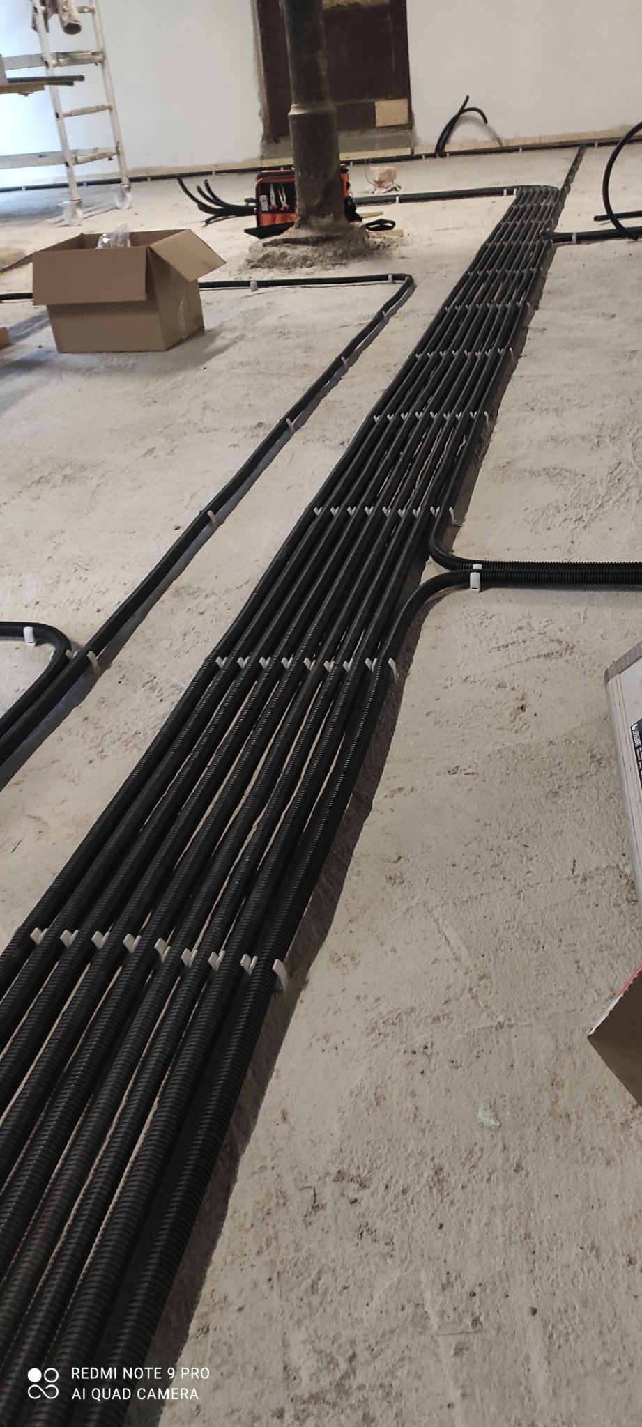 Równolegle ułożone, czarne rury karbowane na surowym betonowym podłożu, przytwierdzone białymi uchwytami, z narzędziami i kartonem w tle, ukazujące etap prac instalacyjnych.