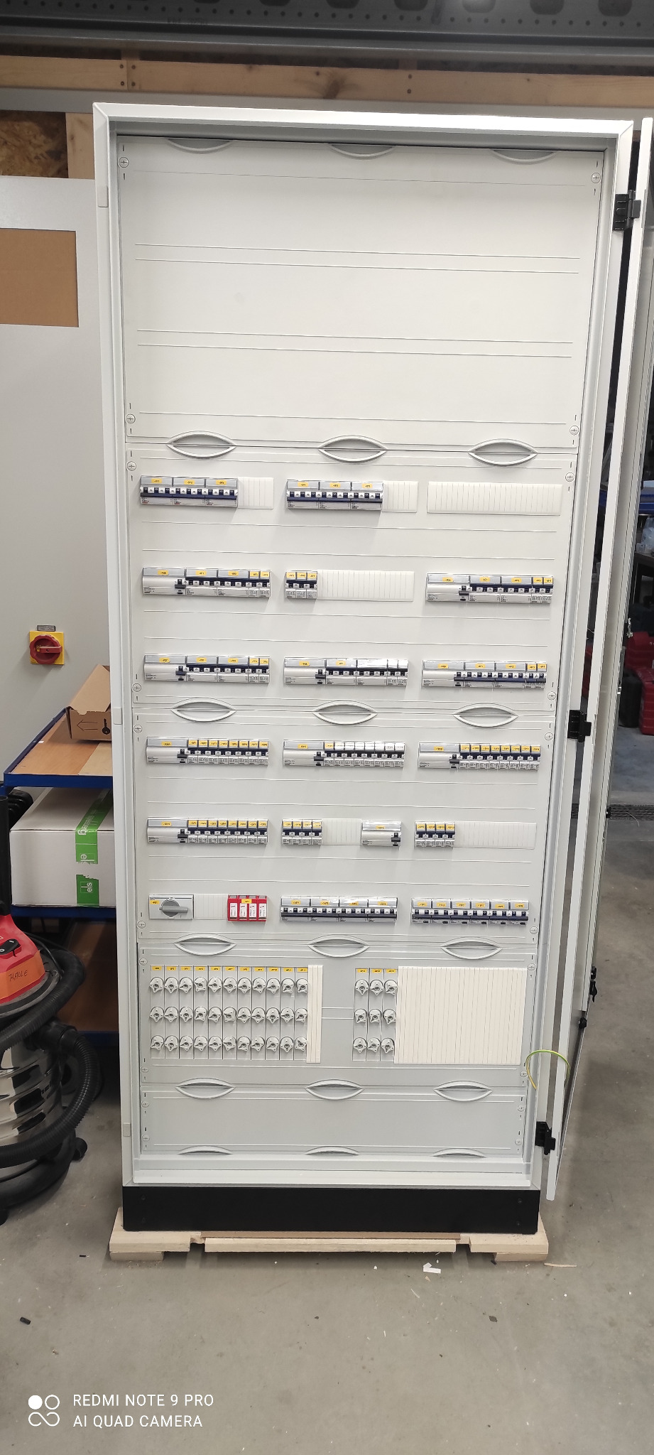 Otwarta, szara szafa elektryczna z widocznymi bezpiecznikami, wyłącznikami i innym osprzętem, umieszczona na drewnianej palecie w pomieszczeniu technicznym.