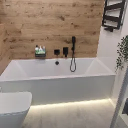 Nowoczesna łazienka z wanną, wiszącą toaletą i drewnopodobnymi panelami ściennymi, oświetlenie LED pod wanną, czarna armatura.