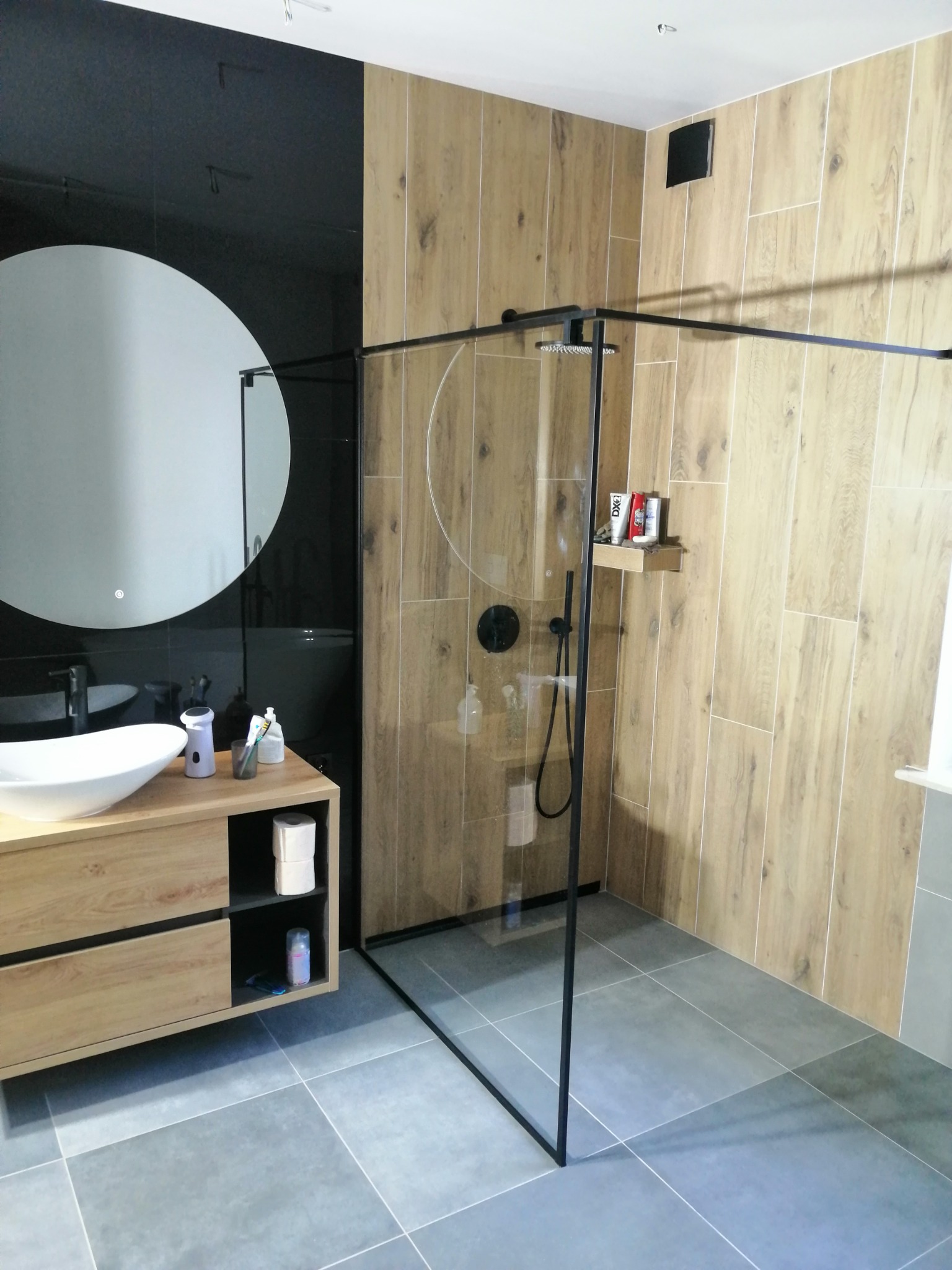 Nowoczesna łazienka z drewnopodobnymi płytkami na ścianach, kabiną prysznicową typu walk-in z czarną armaturą, okrągłym lustrem z podświetleniem i szafką pod umywalką w kolorze jasnego drewna.