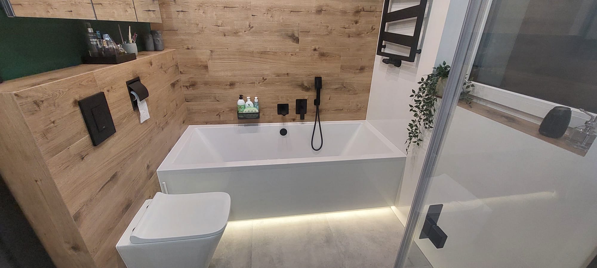 Nowoczesna łazienka z wanną, wiszącą toaletą i drewnopodobnymi panelami ściennymi, oświetlenie LED pod wanną, czarna armatura.