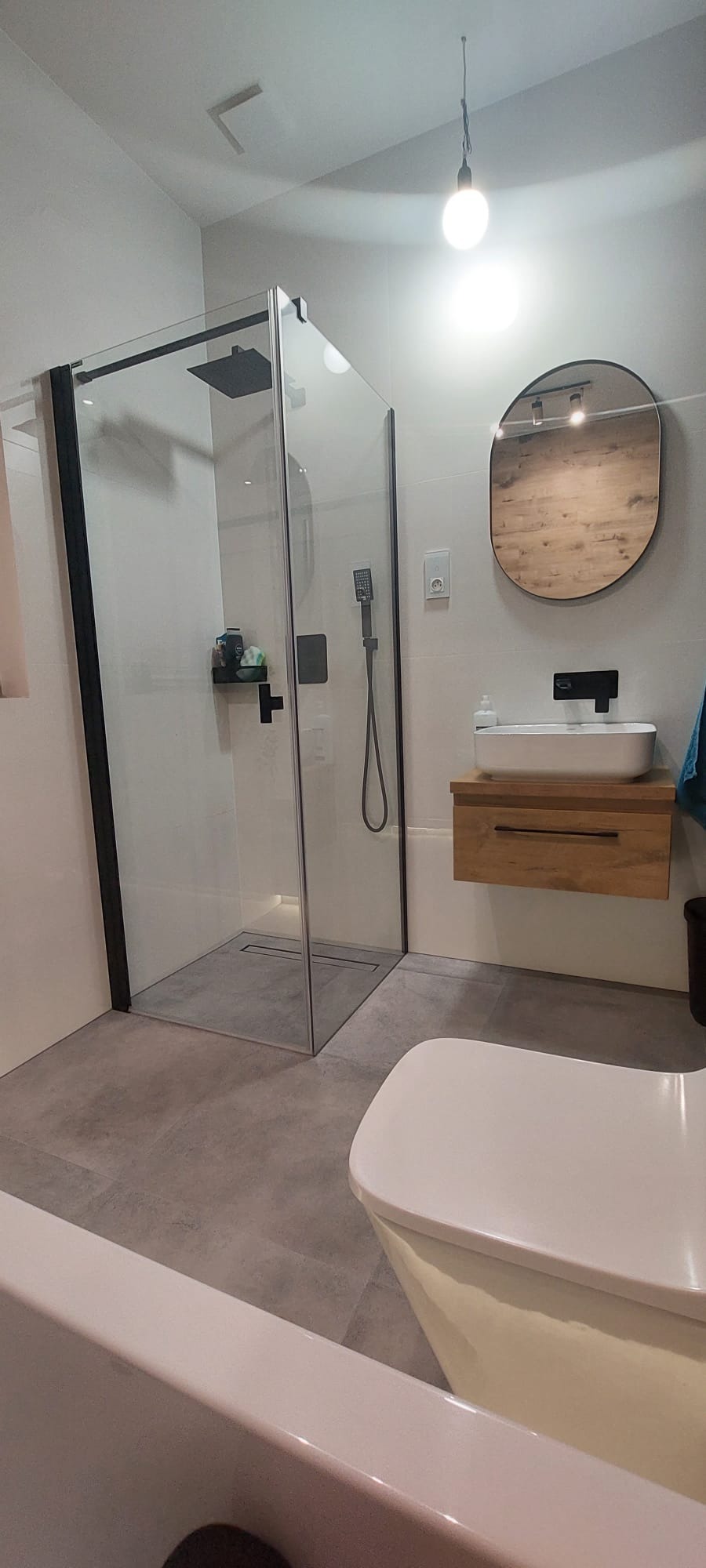 Nowoczesna łazienka z kabiną prysznicową typu walk-in z czarnymi ramami, umywalką nablatową na drewnianej szafce i owalnym lustrem z drewnianą ramą, oświetlona wiszącą żarówką.