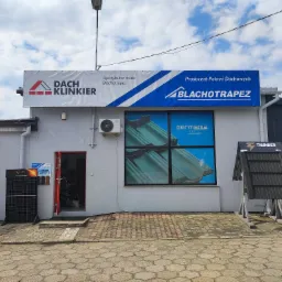 Fasada budynku z szyldami firm Dach Klinkier i Blachotrapez, ekspozycja paneli fotowoltaicznych i próbek pokryć dachowych przed wejściem.