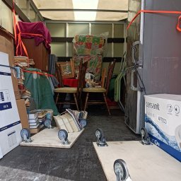 Perfekt Przeprowadzki Sylwester Troszczyński - Wnętrze furgonetki załadowanej meblami, książkami i sprzętem AGD zabezpieczonym pomarańczowymi pasami, część przedmiotów na drewnianych platformach z kółkami.