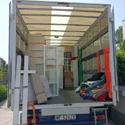 Wnętrze załadowanego busa transportowego z meblami, dywanami i stertą koców, zabezpieczonych pomarańczowymi pasami, widok z zewnątrz na zaparkowany pojazd.