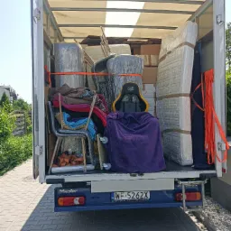 Wnętrze białej furgonetki załadowane do przeprowadzki: meble owinięte folią, kartony, krzesła i fotel gamingowy przykryty fioletowym kocem, zabezpieczone pomarańczowymi pasami.