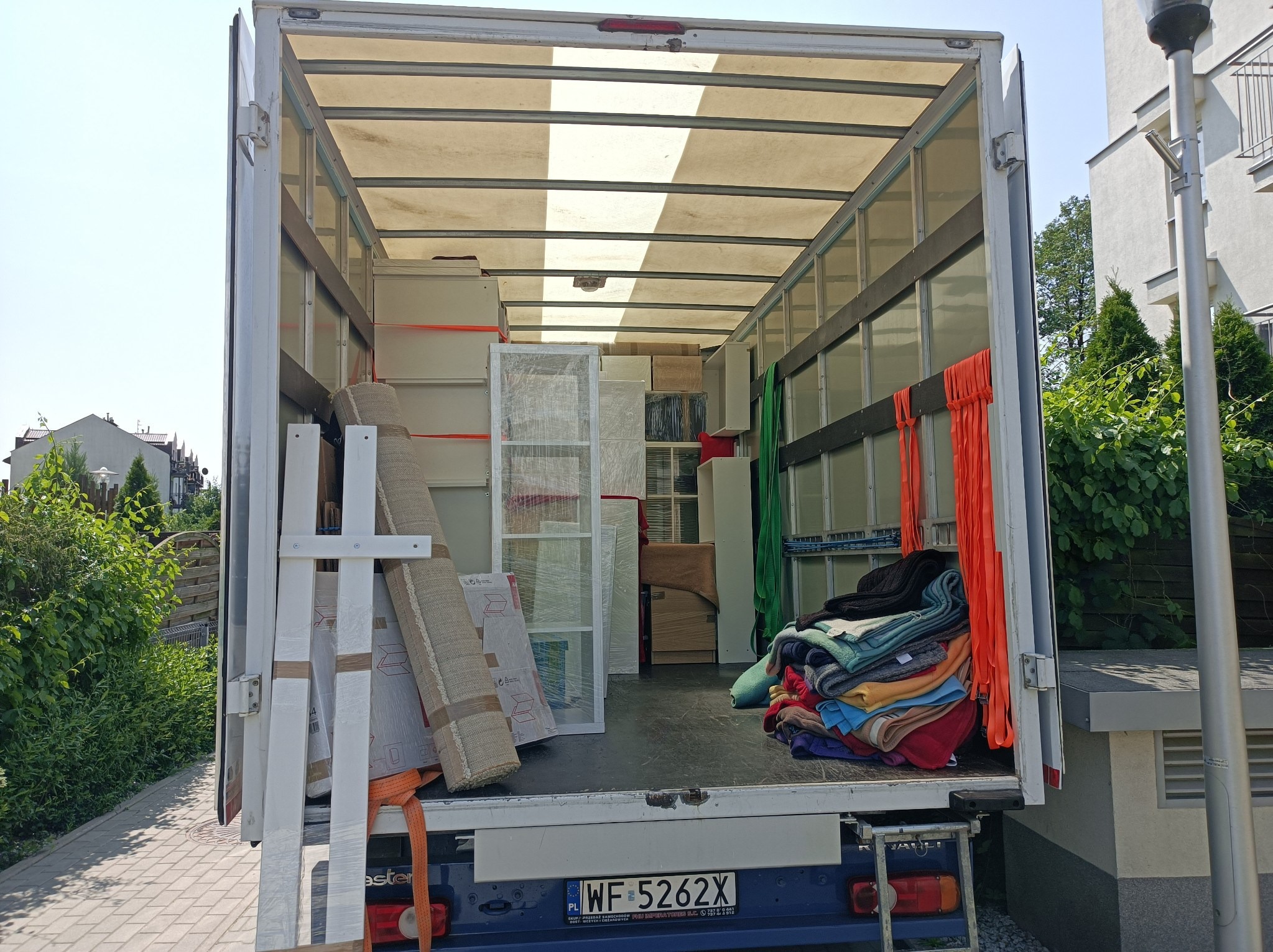 Wnętrze załadowanego busa transportowego z meblami, dywanami i stertą koców, zabezpieczonych pomarańczowymi pasami, widok z zewnątrz na zaparkowany pojazd.