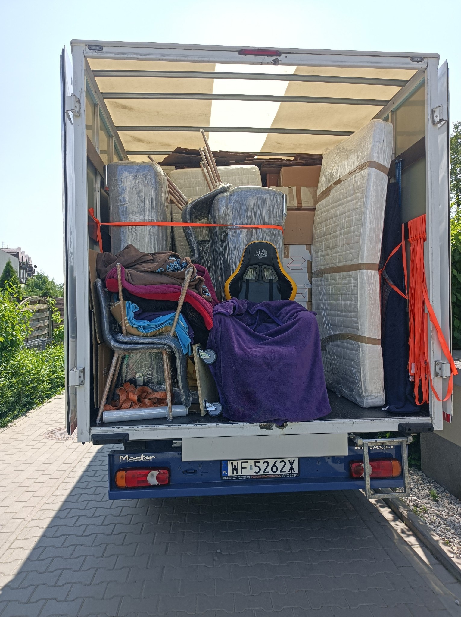 Wnętrze białej furgonetki załadowane do przeprowadzki: meble owinięte folią, kartony, krzesła i fotel gamingowy przykryty fioletowym kocem, zabezpieczone pomarańczowymi pasami.