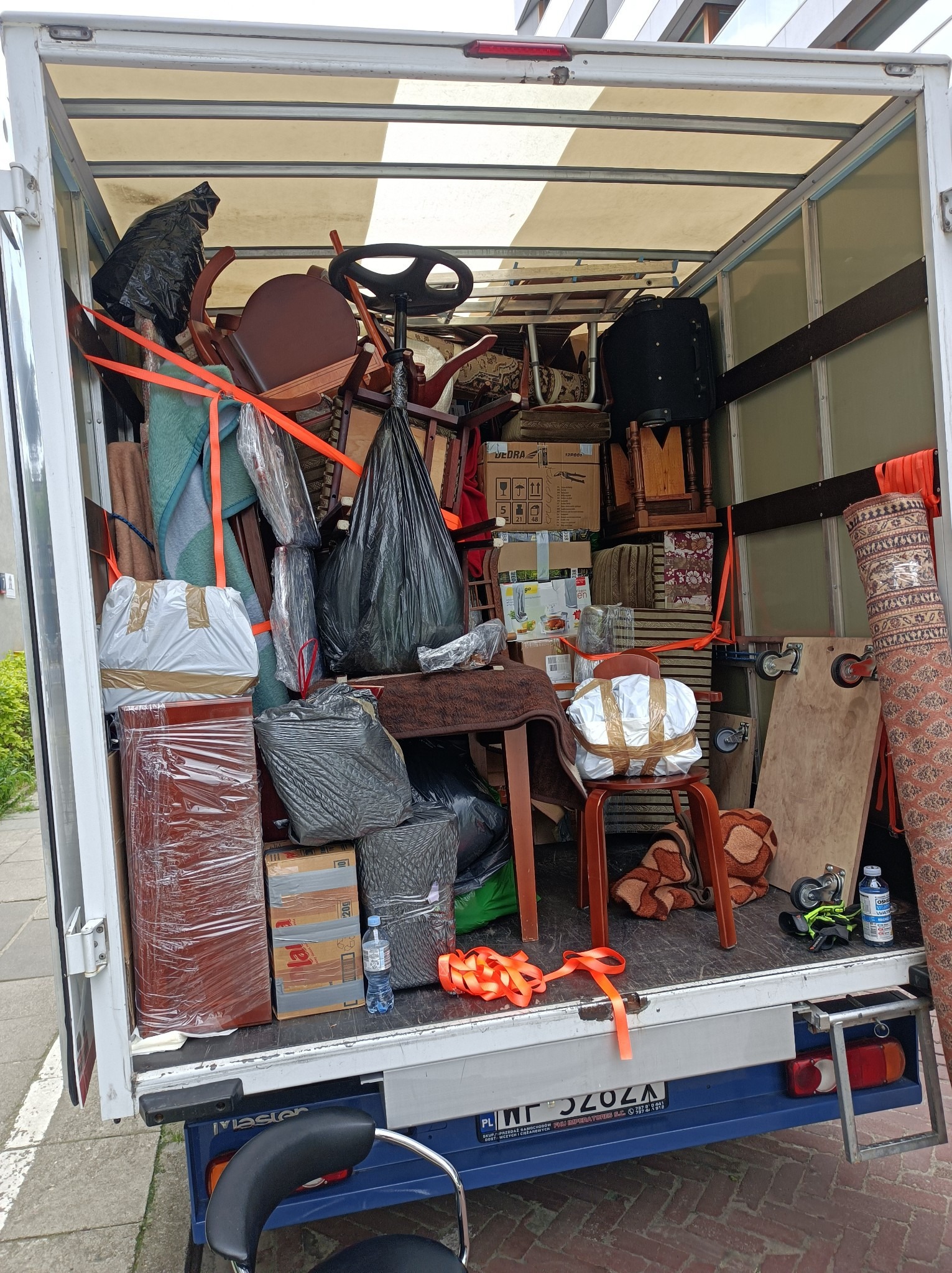 Wnętrze furgonetki wypełnionej meblami, kartonami i workami zabezpieczonymi pomarańczowymi pasami transportowymi podczas relokacji.