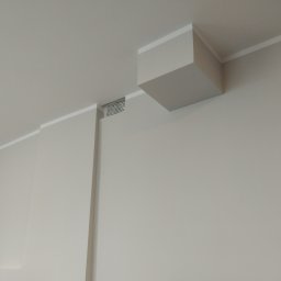 SKBudRem - Minimalistyczne wykończenie wnętrza: gładka, pomalowana na szaro ściana z dekoracyjną listwą przysufitową, kratka wentylacyjna i wystający fragment sufitu w kształcie sześcianu.