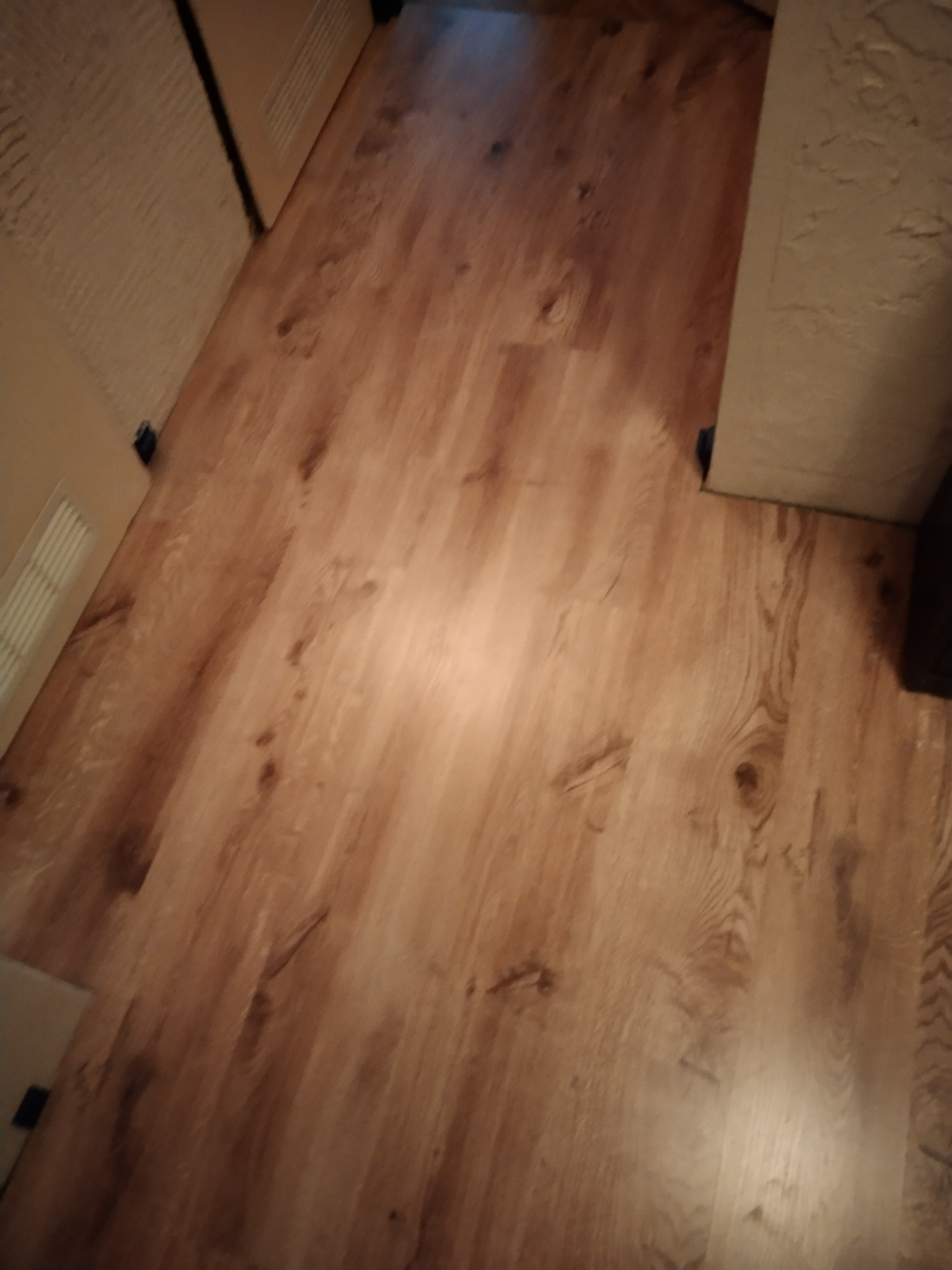 Ułożone panele podłogowe z widocznymi detalami tekstury drewna, częściowo oświetlone, z fragmentem białej ściany i drzwi w tle.