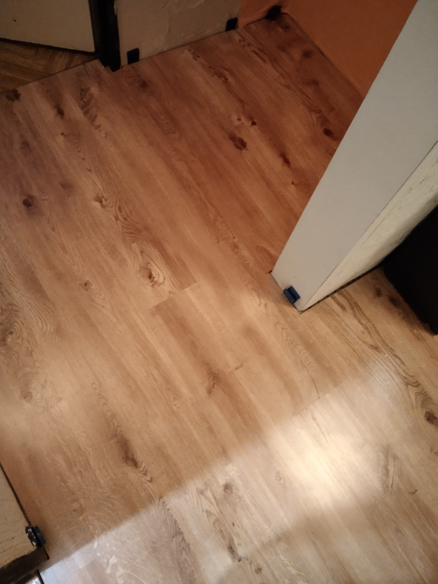 Fragment podłogi wyłożonej panelami imitującymi drewno, widoczne niebieskie kliny dystansowe przy ścianie i futrynie, w trakcie montażu.