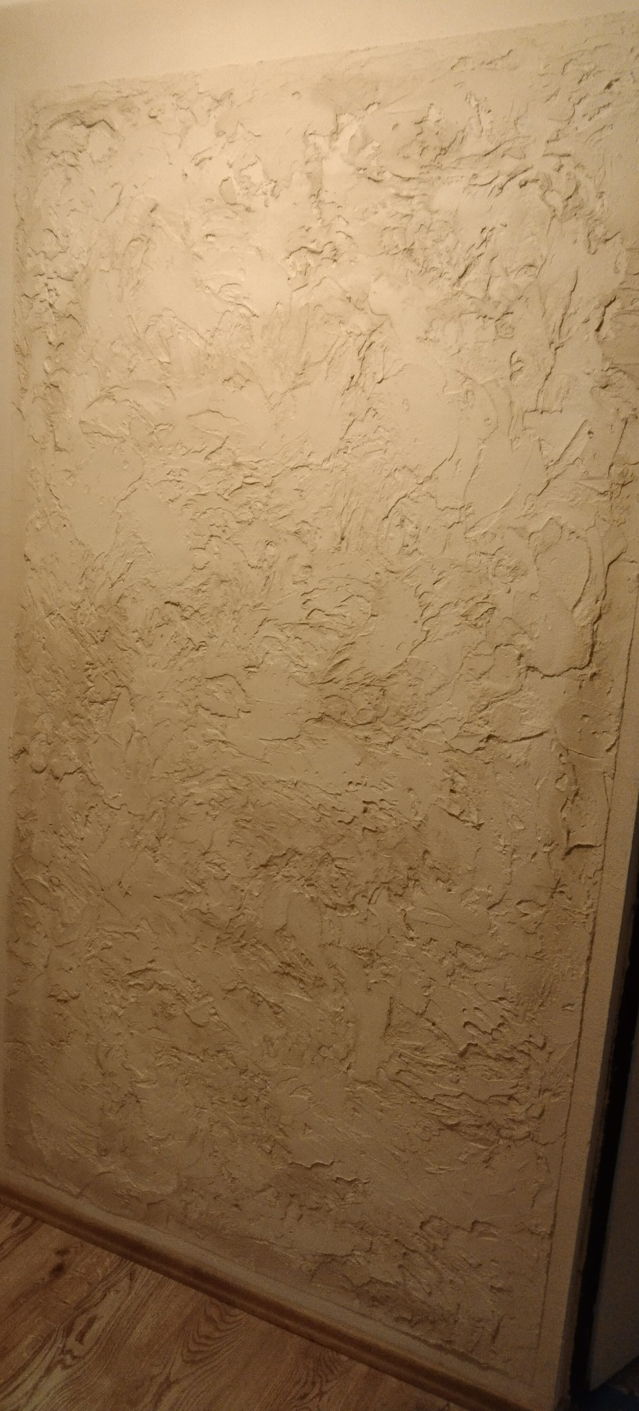 Beżowa ściana z wyraźną, nieregularną fakturą, imitującą kamień lub piasek, oświetlona z góry, widoczne fragmenty drewnianej podłogi i listwy przypodłogowej.