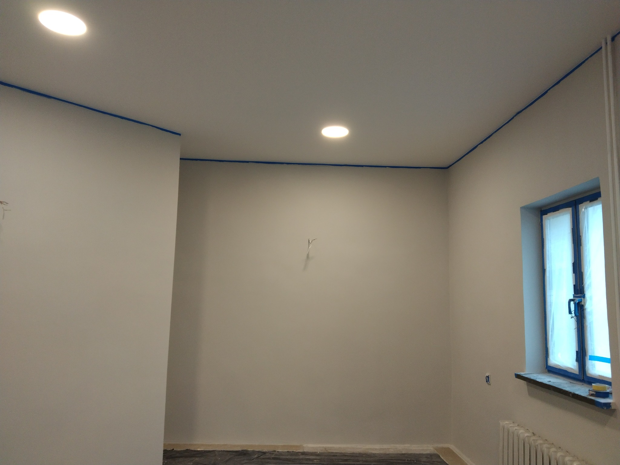 Pokój przygotowany do malowania: ściany i sufit zabezpieczone taśmą malarską, okno zakryte folią ochronną, podłoga przykryta folią malarską, widoczne dwa wbudowane punkty świetlne w suficie.