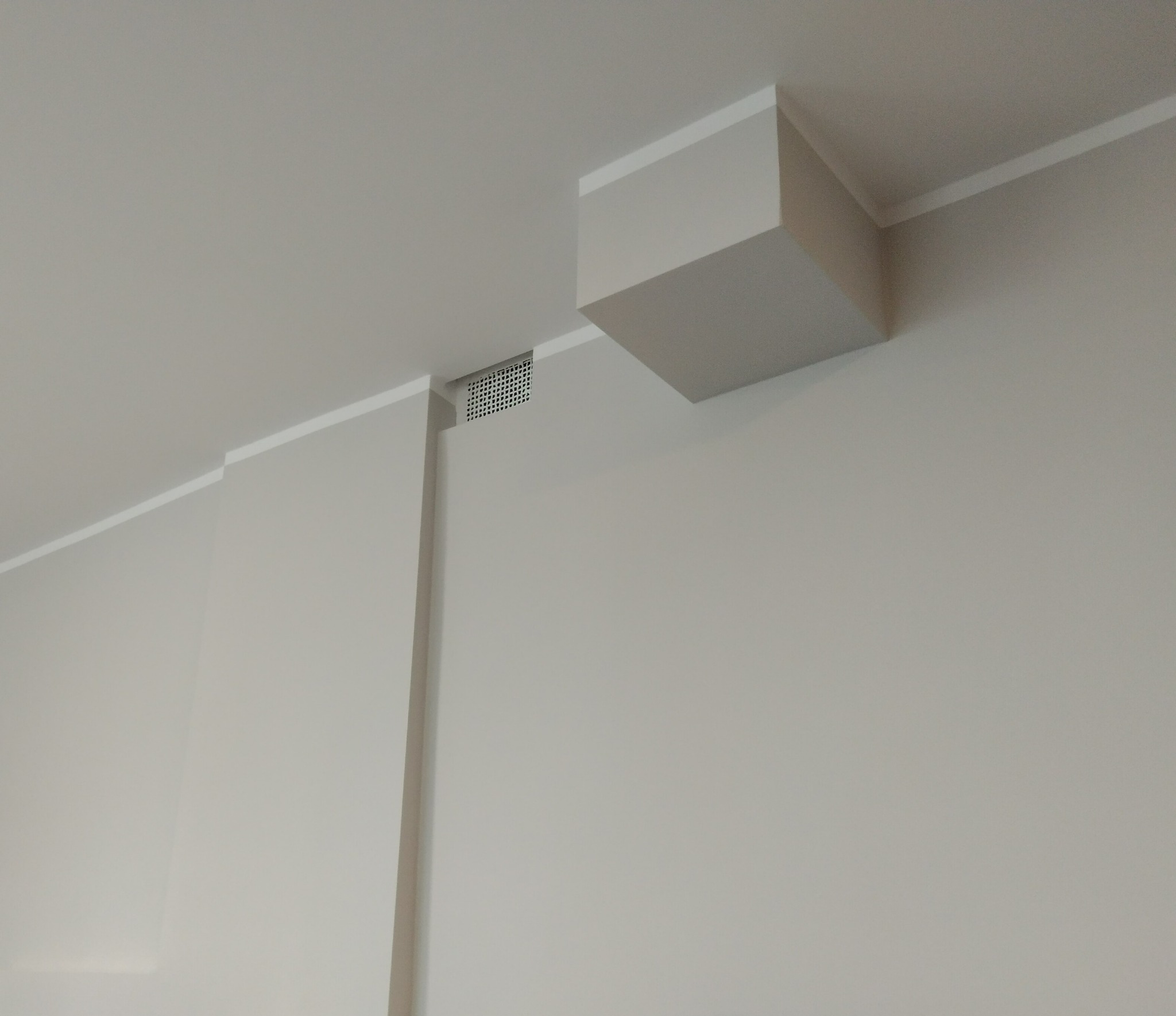 Minimalistyczne wykończenie wnętrza: gładka, pomalowana na szaro ściana z dekoracyjną listwą przysufitową, kratka wentylacyjna i wystający fragment sufitu w kształcie sześcianu.