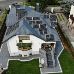 Widok z góry na dom z szarym dachem, na którym zamontowano panele słoneczne; widoczny ogród z elementami dekoracyjnymi i podjazd do garażu.