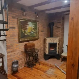 Wnętrze pokoju z ceglaną ścianą, spiralnymi schodami, kominkiem kaflowym, obrazem w złotej ramie, starą maszyną do szycia i mężczyzną w tle.