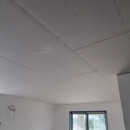 Sufit z płyt kartonowo-gipsowych, widoczne łączenia płyt i wkręty, surowe wnętrze z oknami, instalacja elektryczna w trakcie montażu.