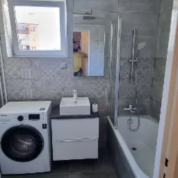 Wyremontowana łazienka z szarymi płytkami, białą pralką Samsung, umywalką na szafce i wanną z parawanem, widok zza otwartych drzwi.