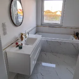 Nowoczesna łazienka z białymi płytkami imitującymi marmur, wisząca szafka pod umywalką z złotą armaturą, okrągłe lustro na skórzanym pasku i złote oświetlenie.