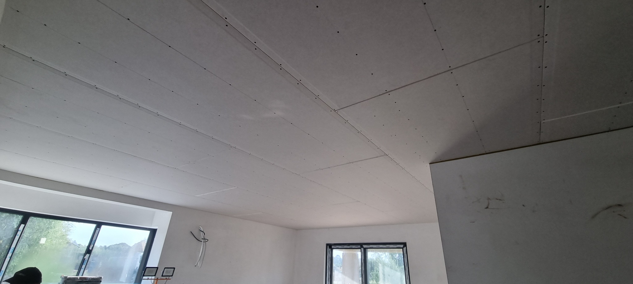 Sufit z płyt kartonowo-gipsowych, widoczne łączenia płyt i wkręty, surowe wnętrze z oknami, instalacja elektryczna w trakcie montażu.