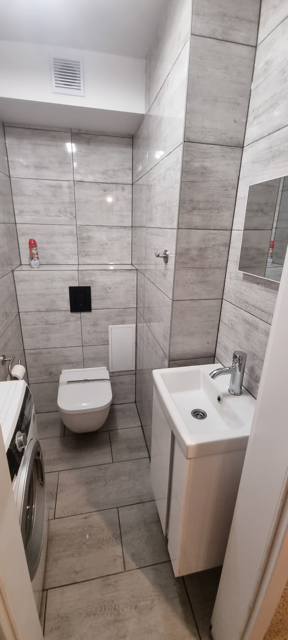 Wykończona łazienka w szarościach z podwieszaną toaletą, umywalką z szafką, pralką i grzejnikiem drabinkowym, widok z poziomu podłogi