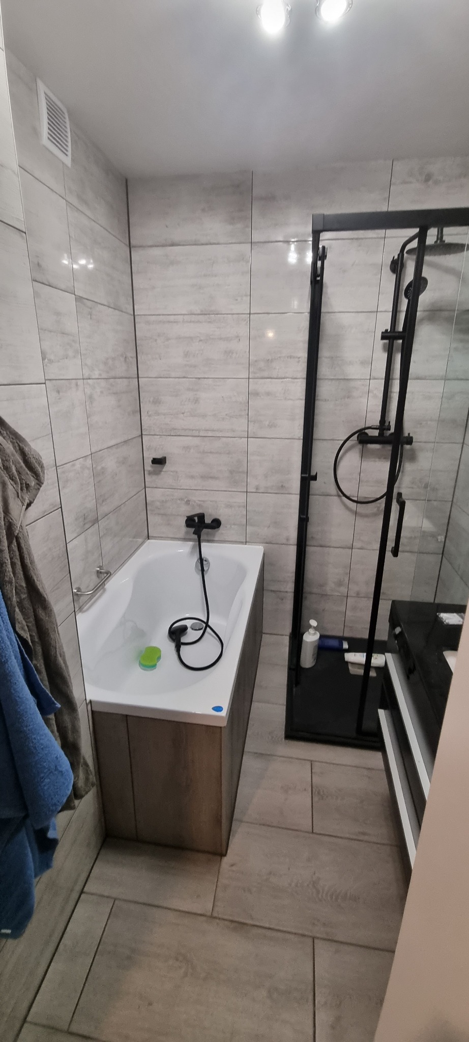 Nowoczesna łazienka po remoncie z wanną obudowaną drewnopodobną okładziną, czarną armaturą i kabiną prysznicową typu walk-in z czarnymi profilami.