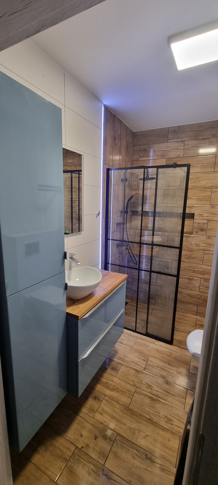 Nowoczesna łazienka z drewnopodobnymi płytkami, kabiną prysznicową w czarnej ramie i szafką umywalkową w odcieniach błękitu z drewnianym blatem.