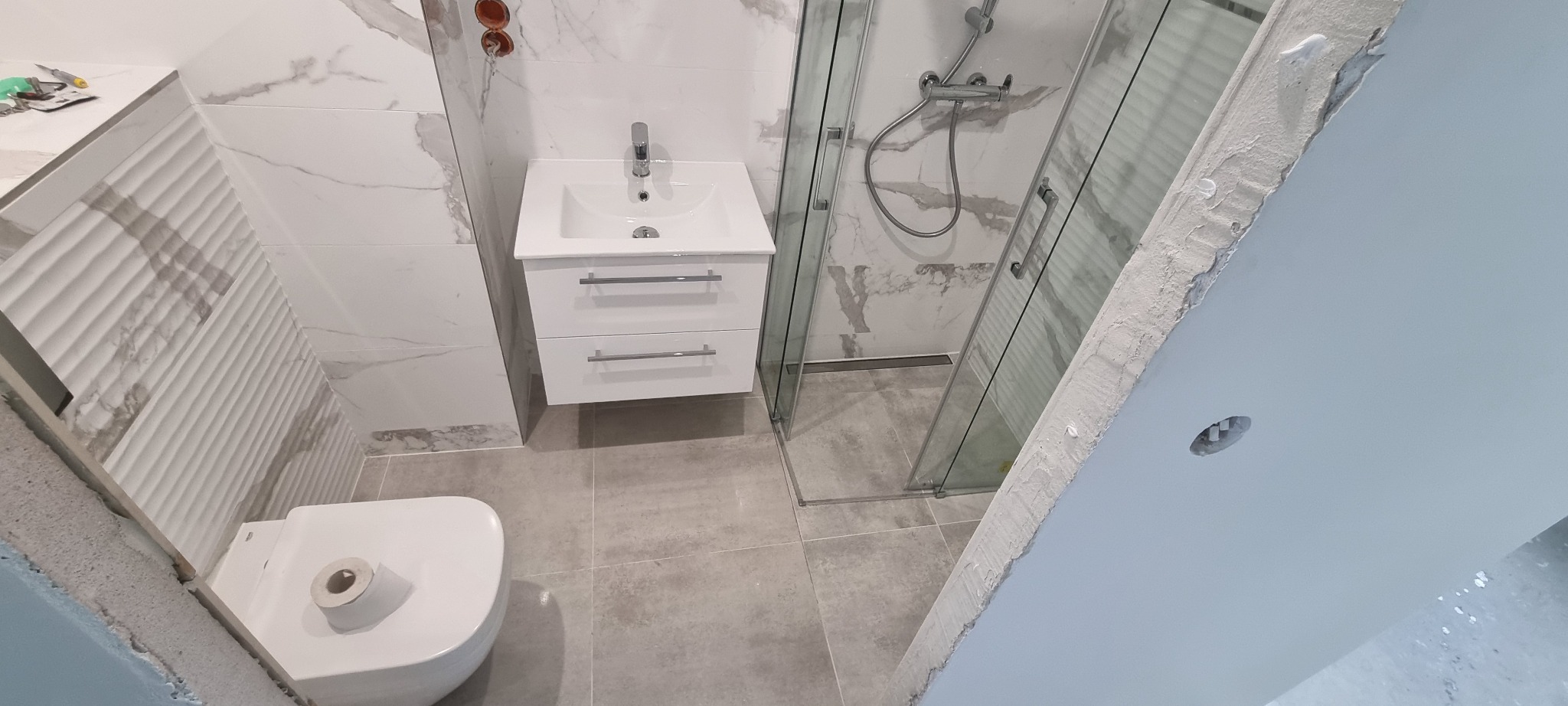 Nowoczesna łazienka w trakcie wykańczania: biała umywalka z szafką, toaleta z rolką papieru, kabina prysznicowa z odpływem liniowym, płytki imitujące marmur na ścianach, szare płytki podłogowe...